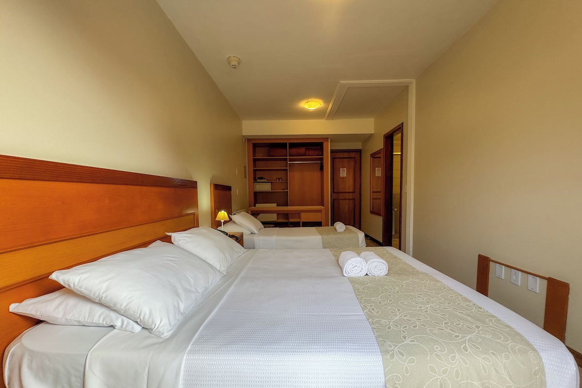 A hotel room with two beds, a wooden headboard, a bedside table with a lamp, and a wardrobe.