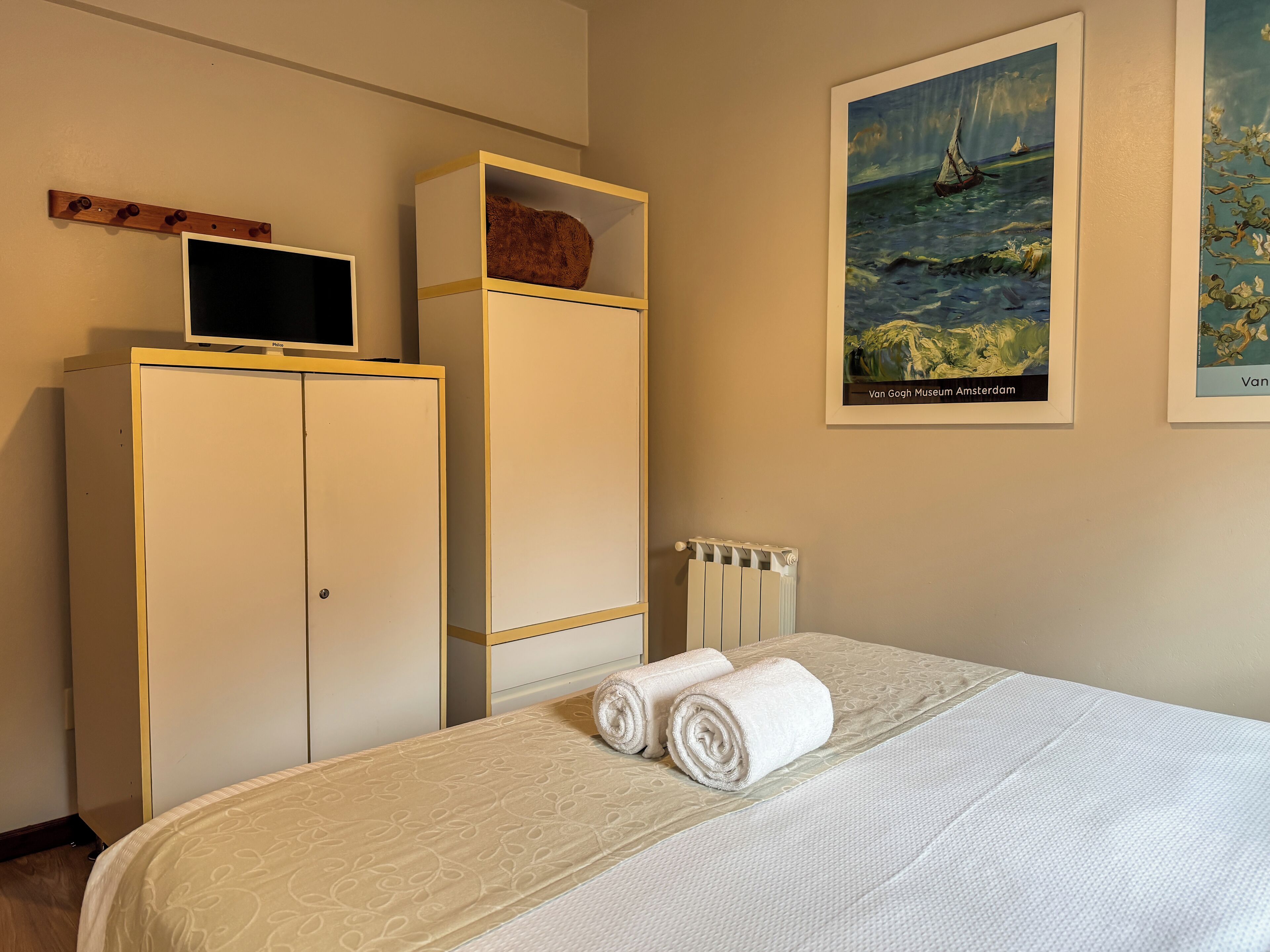 Una habitación de hotel con cama, armario, radiador y un cuadro de un velero.