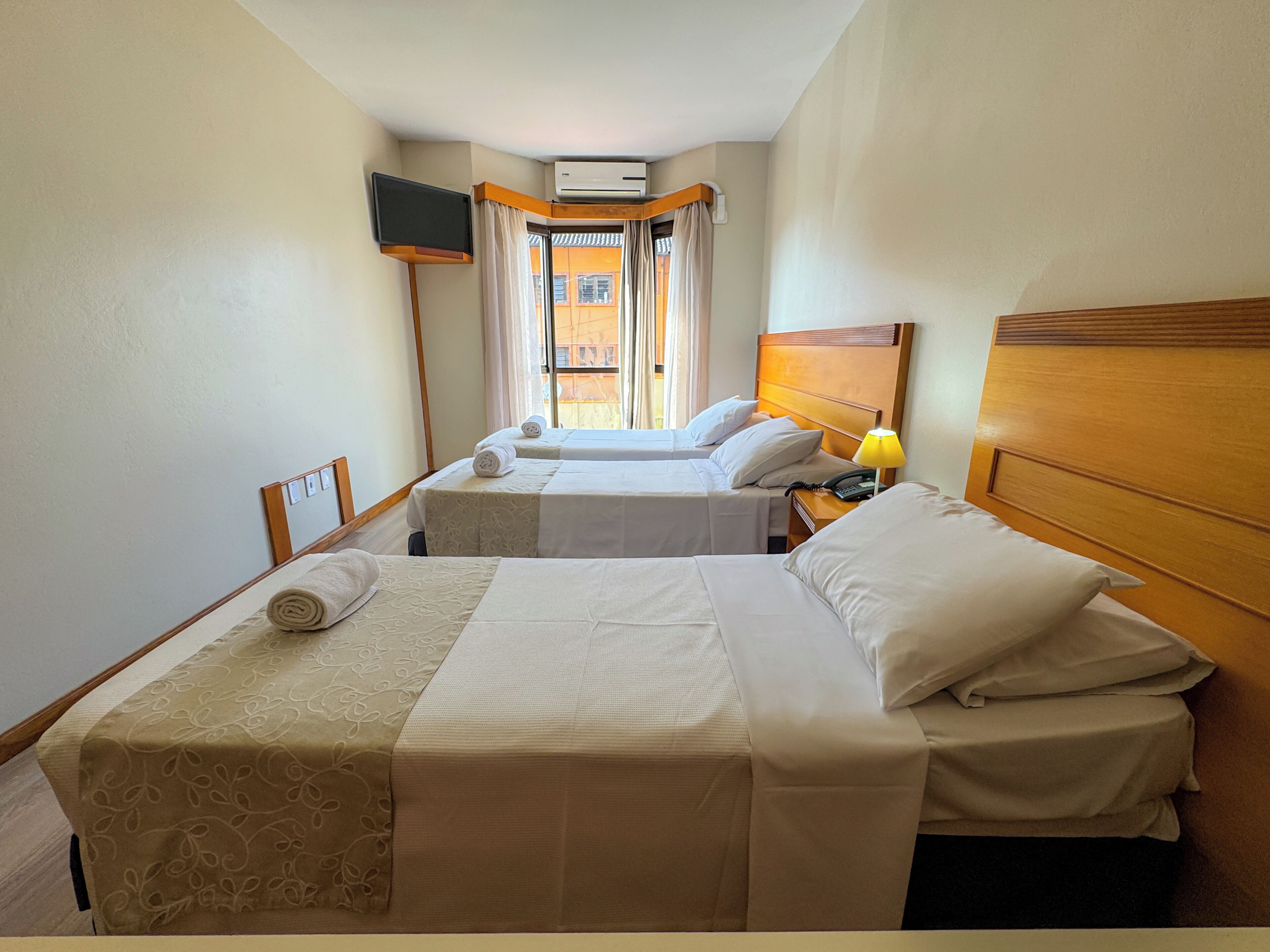 A hotel room with two beds, a TV mounted on the wall, an air conditioning unit, and a view of the outside.