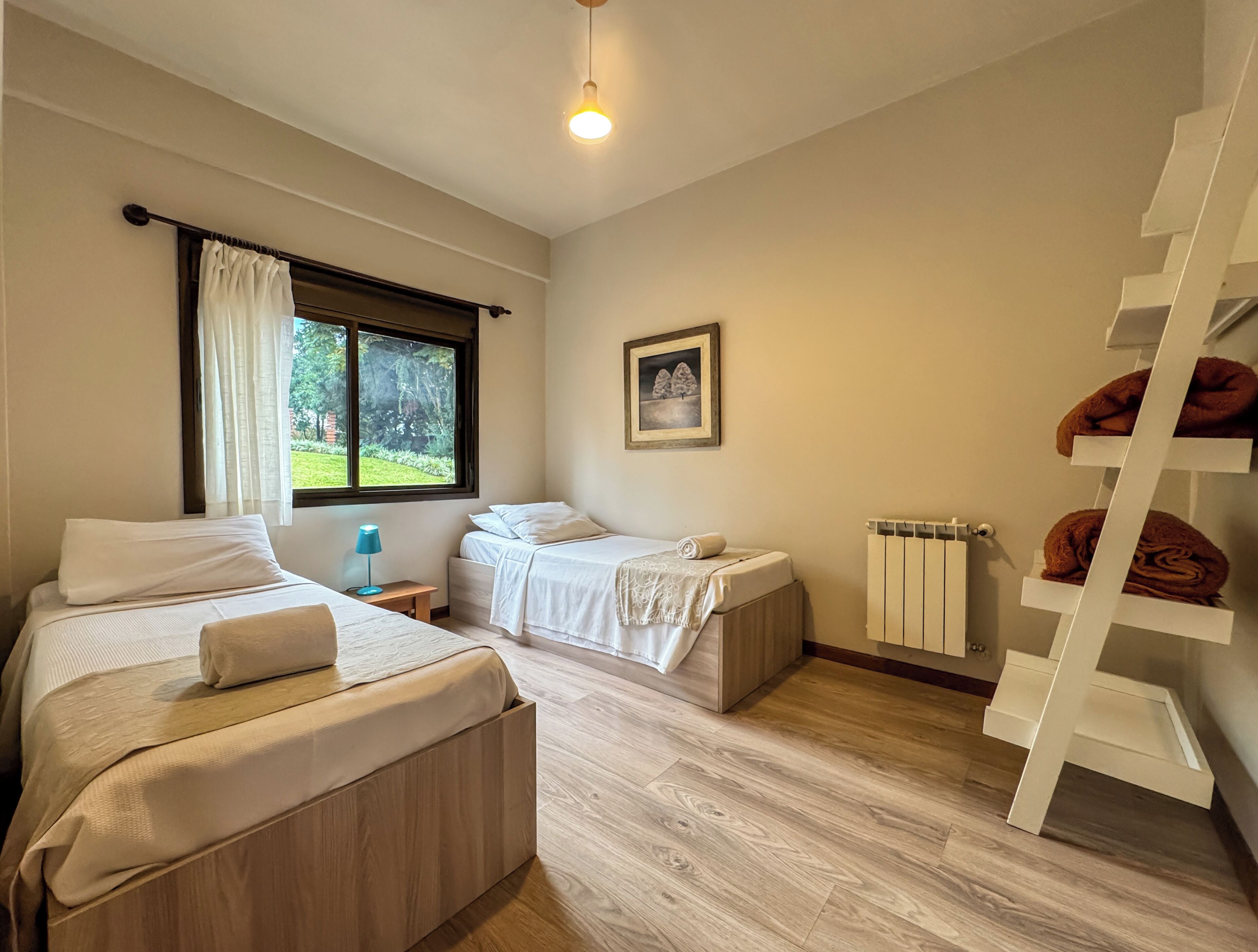 A bedroom with two beds, a window with curtains, a bedside table, a radiator, and a wooden floor.