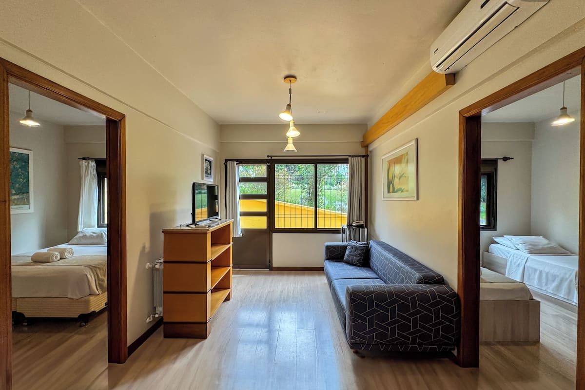 A hotel room with a bed, a sofa, a desk with a computer, a wardrobe, and a window with a view of a yellow building and greenery.