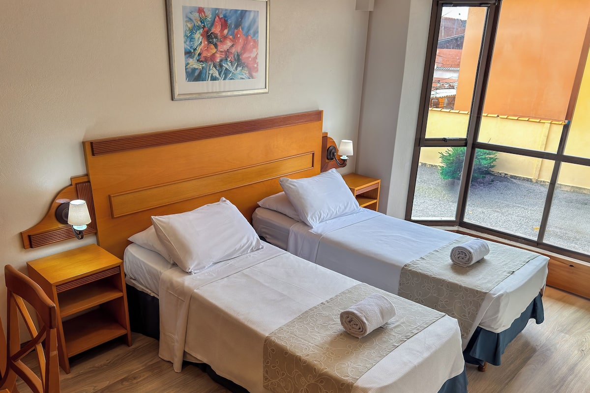 A hotel room with two single beds, wooden headboards, a nightstand with a lamp, a small table, and a window with a view of the outside.