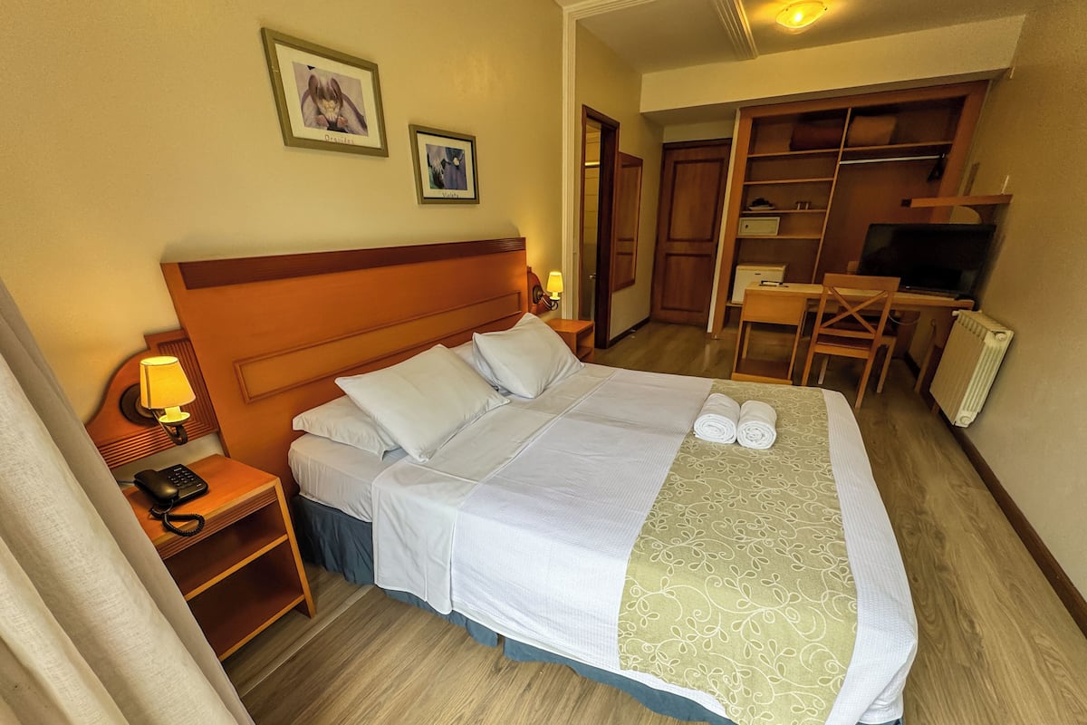 A hotel room with a large bed, a desk, a chair, a wardrobe, and two framed pictures on the wall.