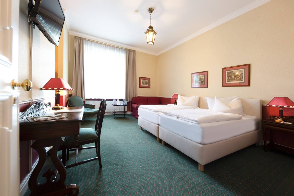 A hotel room with a bed, a desk with a lamp, a chair, a small table, and a window with curtains.