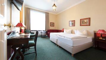 Une chambre d’hôtel avec un lit, un bureau avec une lampe, une chaise, une petite table et une fenêtre avec des rideaux.