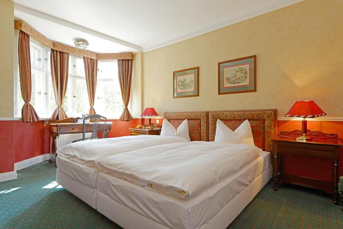 A double bed in a hotel room with two framed pictures on the wall, a desk with a lamp, and a window with curtains.
