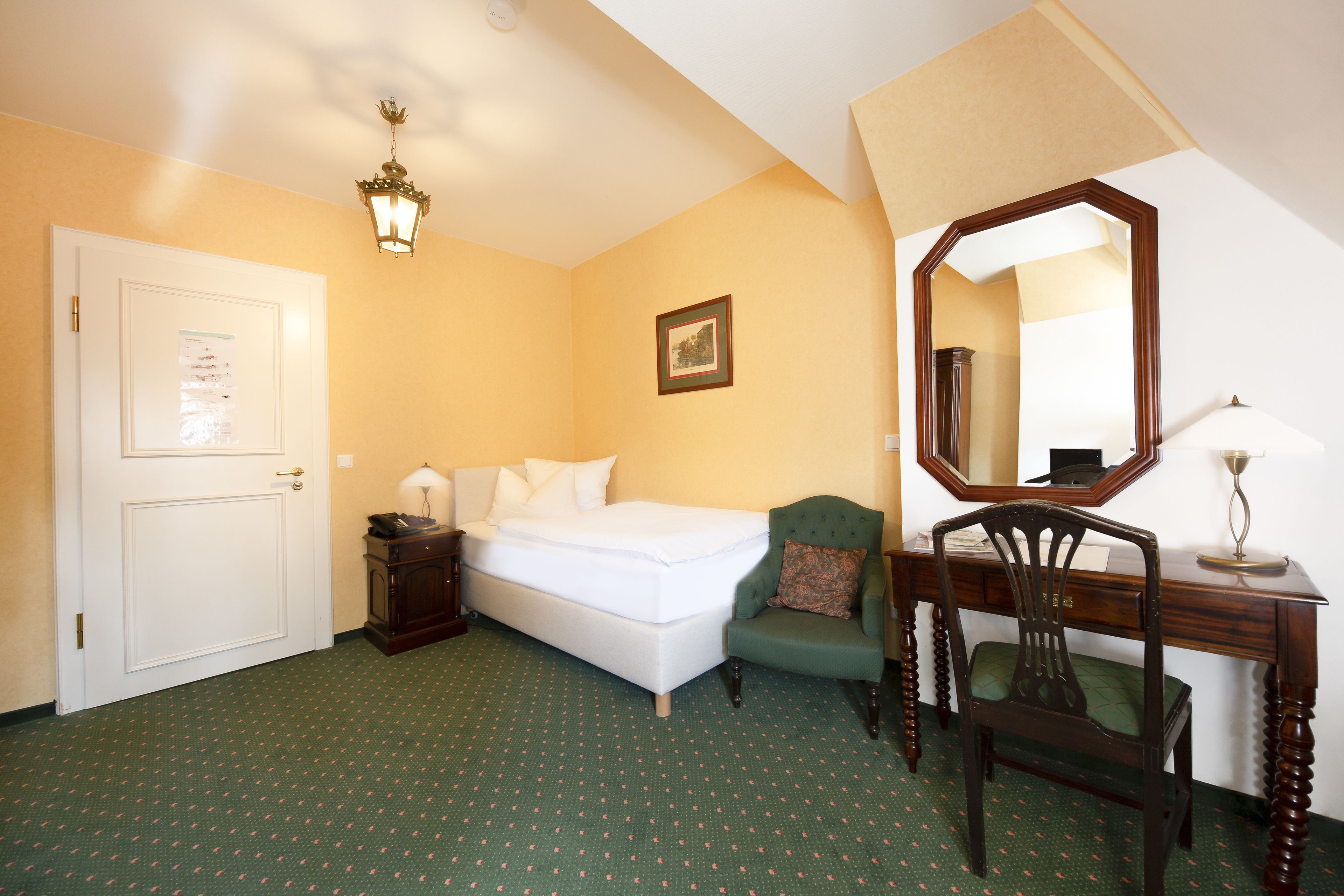 A hotel room with a bed, a desk with a mirror, a chair, a lamp, and a door.
