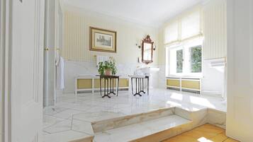 Una habitación luminosa con suelo de mármol, un banco blanco, una mesa con una planta y una ventana con persianas.