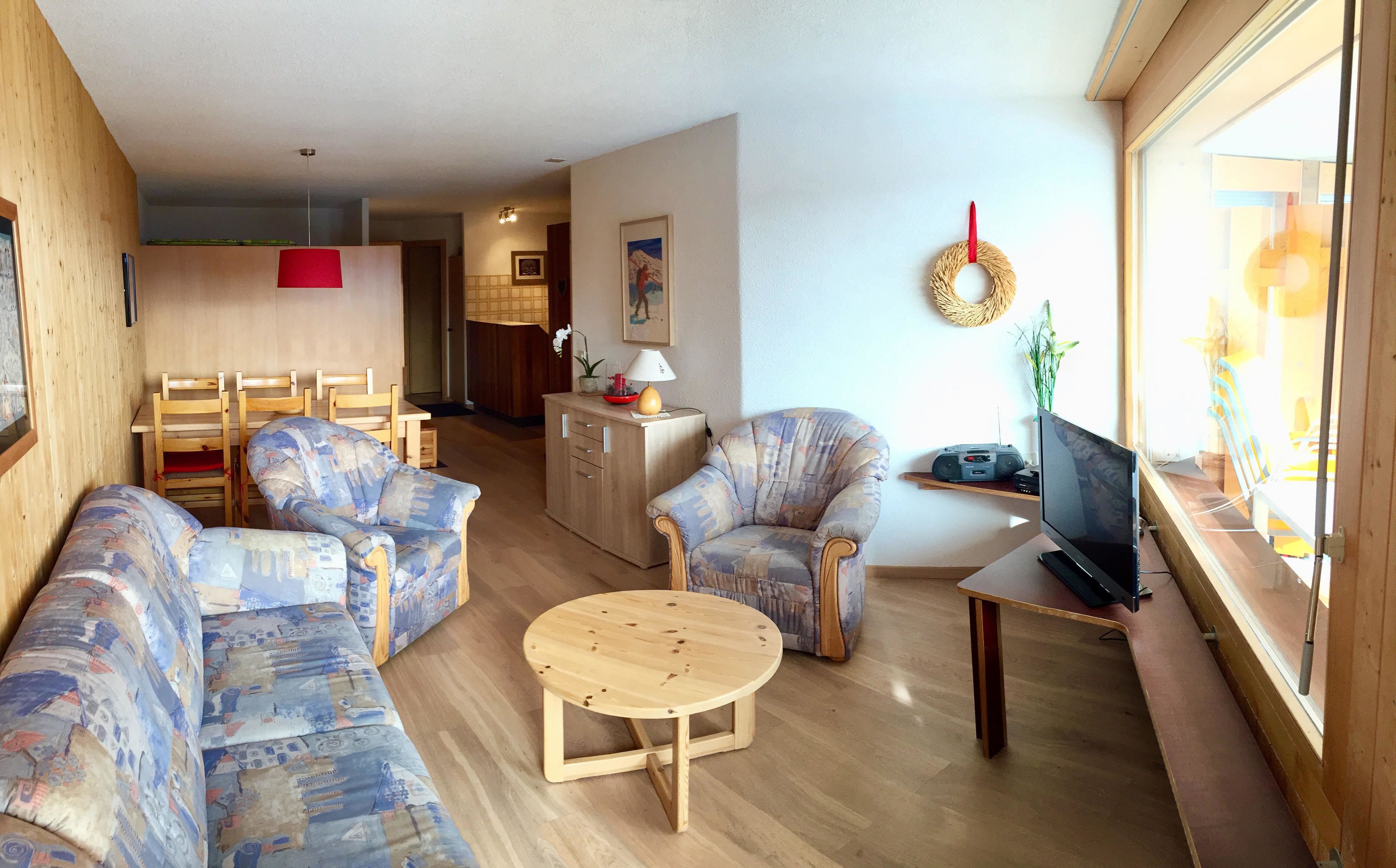 Ein Wohnzimmer mit einer Sofagarnitur, einem Couchtisch, einem Fernseher und einem Essbereich im Hintergrund.