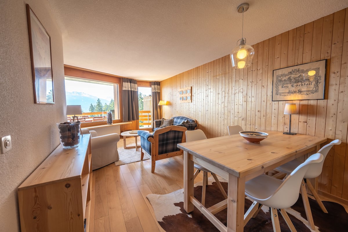 A wooden-paneled room with a dining table, chairs, a sofa, and a view of mountains through a window.