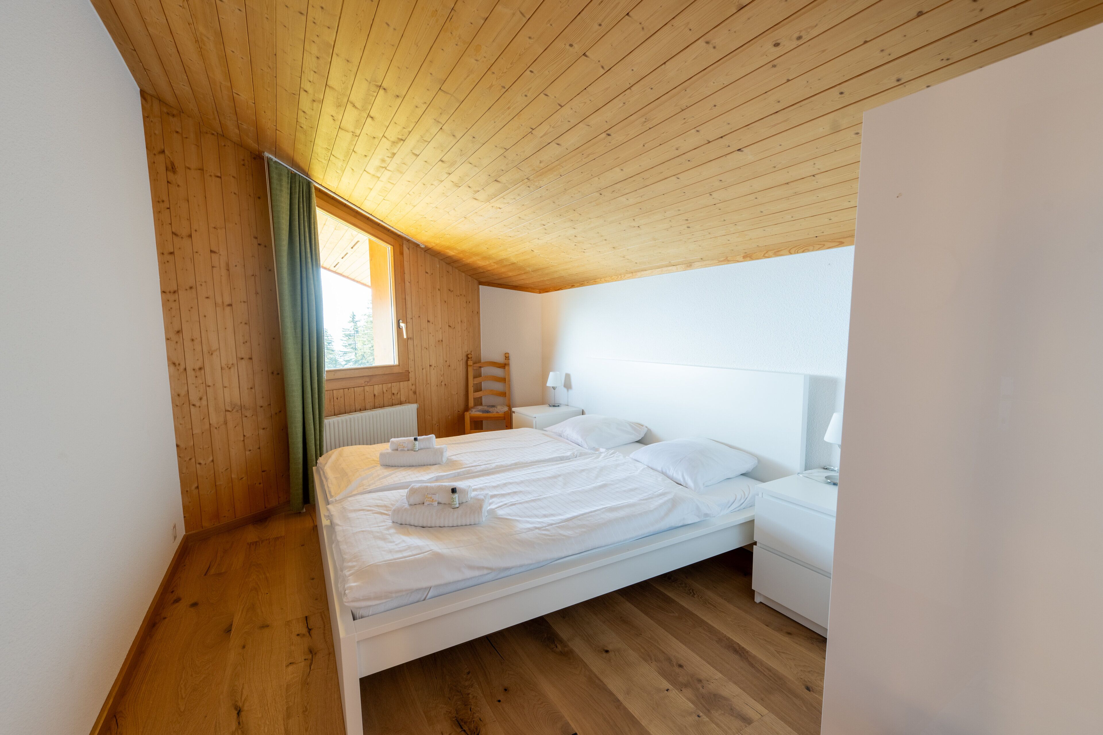 A bedroom with a wooden ceiling and sloped roof, two beds with white linens, a nightstand, and a window with a view of trees.