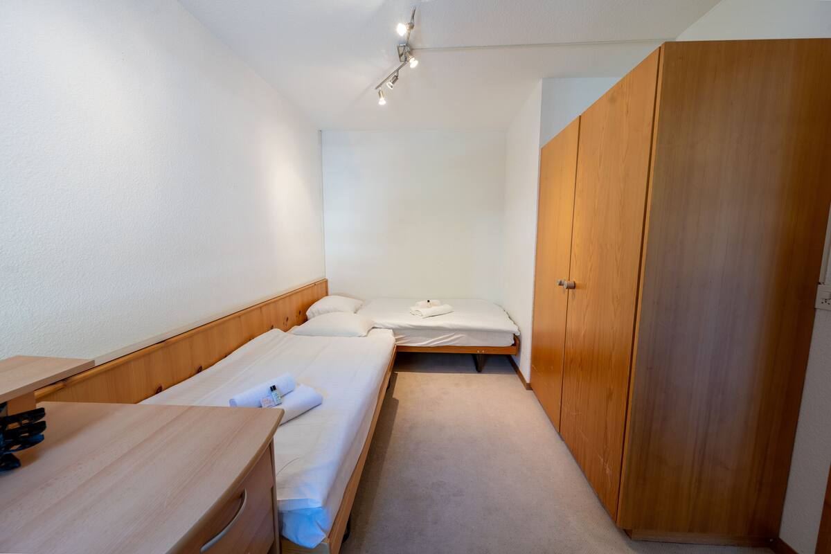 A small, compact room with two single beds, a wooden wardrobe, a desk with a chair, and a ceiling light fixture.