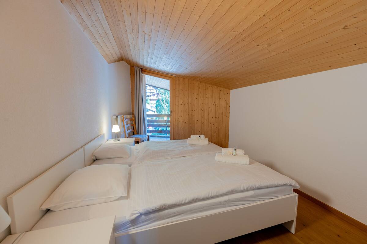A bedroom with a wooden ceiling, a large bed with white bedding, a nightstand, and a window with a view of buildings.