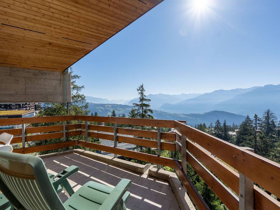 Un terrazzo in legno con sedie verdi e un tavolo, con vista su un paesaggio montuoso e cielo sereno.