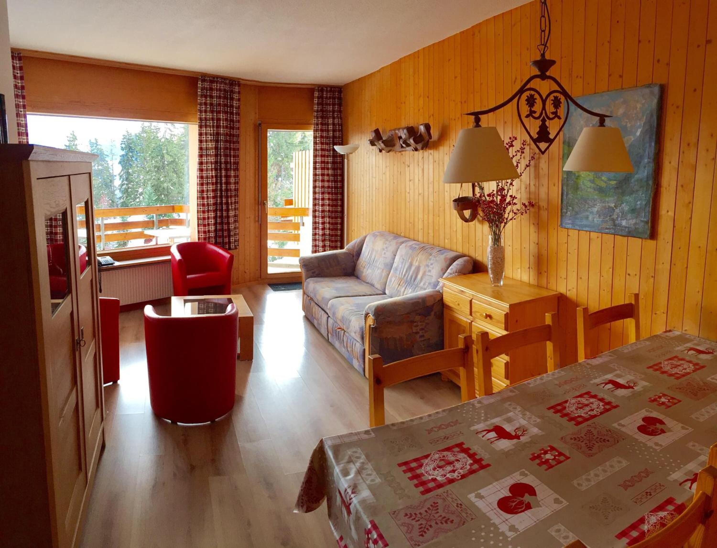 A cozy room with a sofa, a dining table, and a red chair. There is a window with curtains, a lamp, and a painting on the wall.