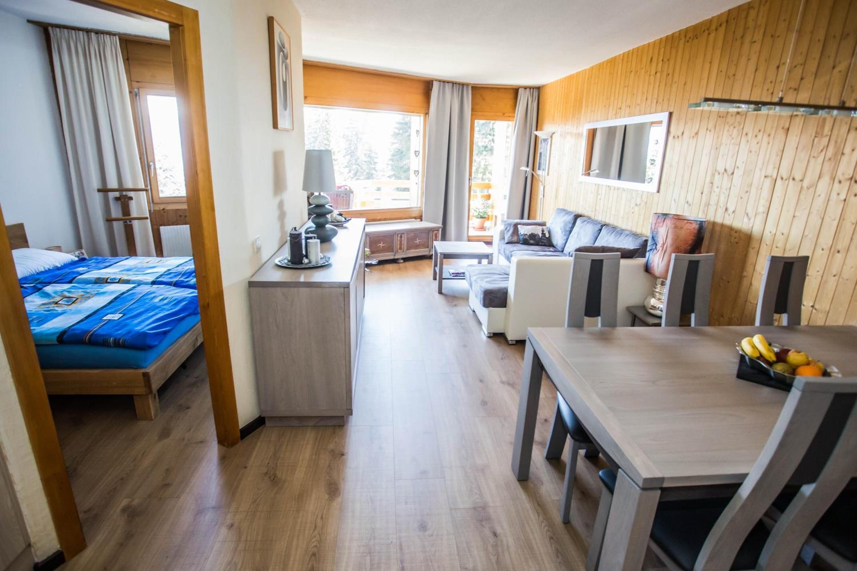 A modern hotel room with a wooden interior, a bed, a desk, a sofa, and a dining table with fruit.