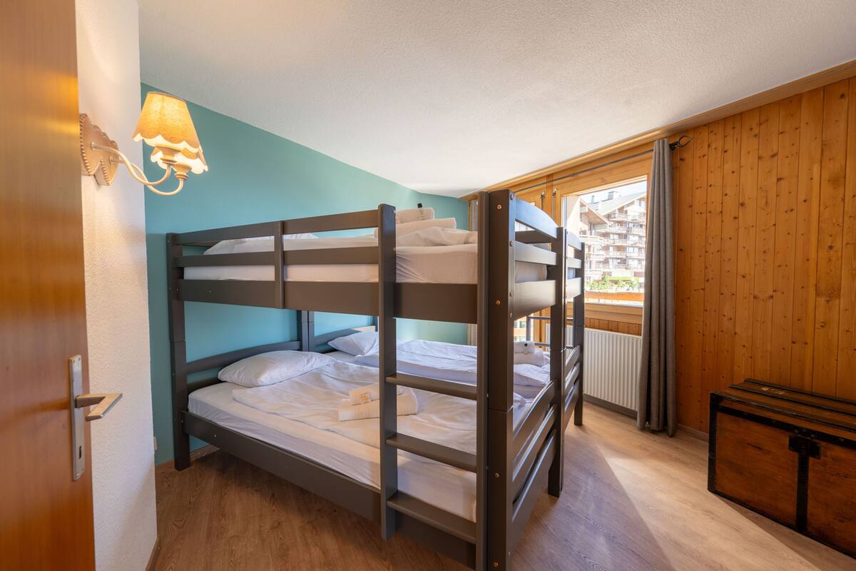 A bunk bed room with a wooden headboard, a bedside lamp, and a view of a building through a window.