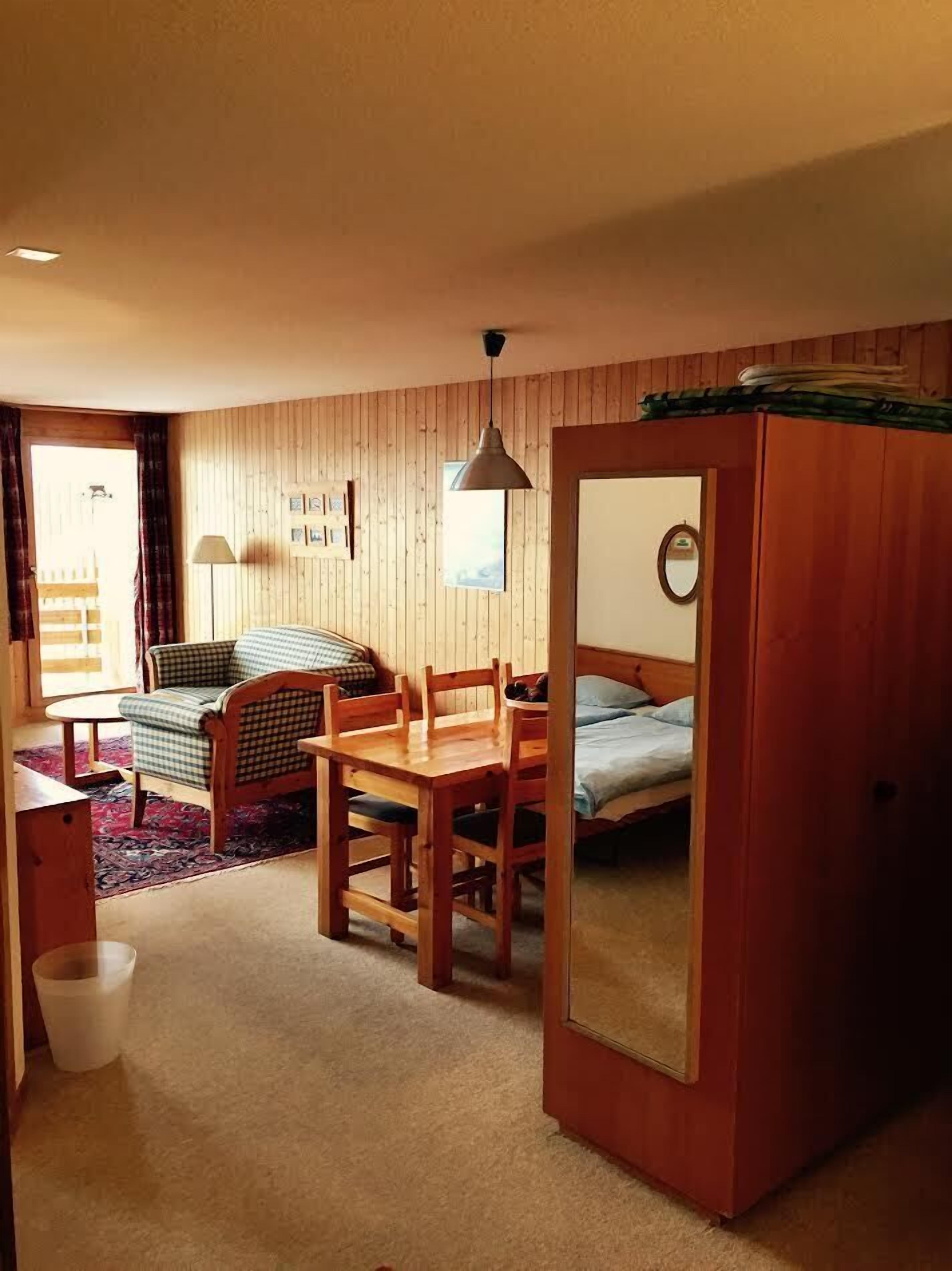 A cozy room with a wooden bed, a small table, a chair, and a desk. There is a mirror on the wall and a window with curtains.