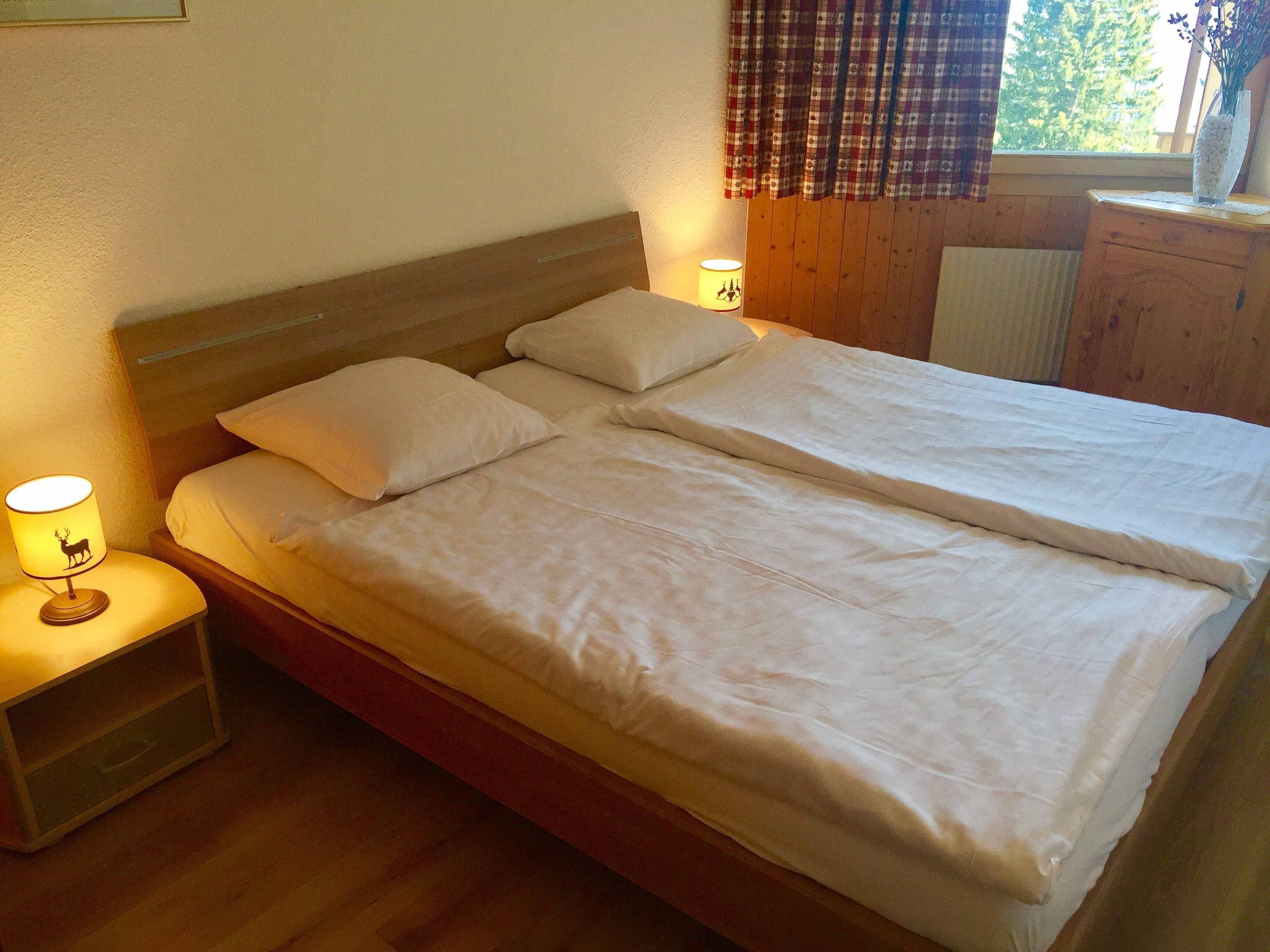 Uma cama de casal com lençóis brancos, uma mesa de cabeceira com abajur e uma janela com cortinas xadrez.