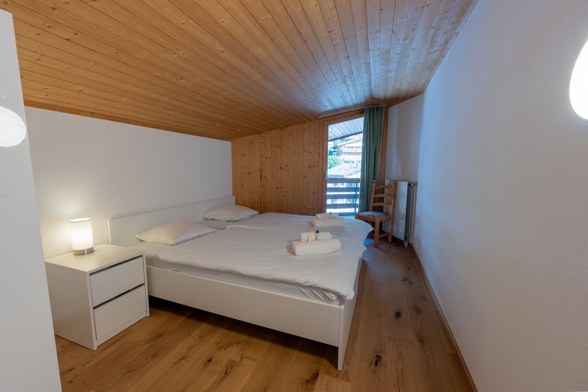 A bedroom with a wooden ceiling, a bed with white linens, a nightstand, and a window with a view of buildings outside.
