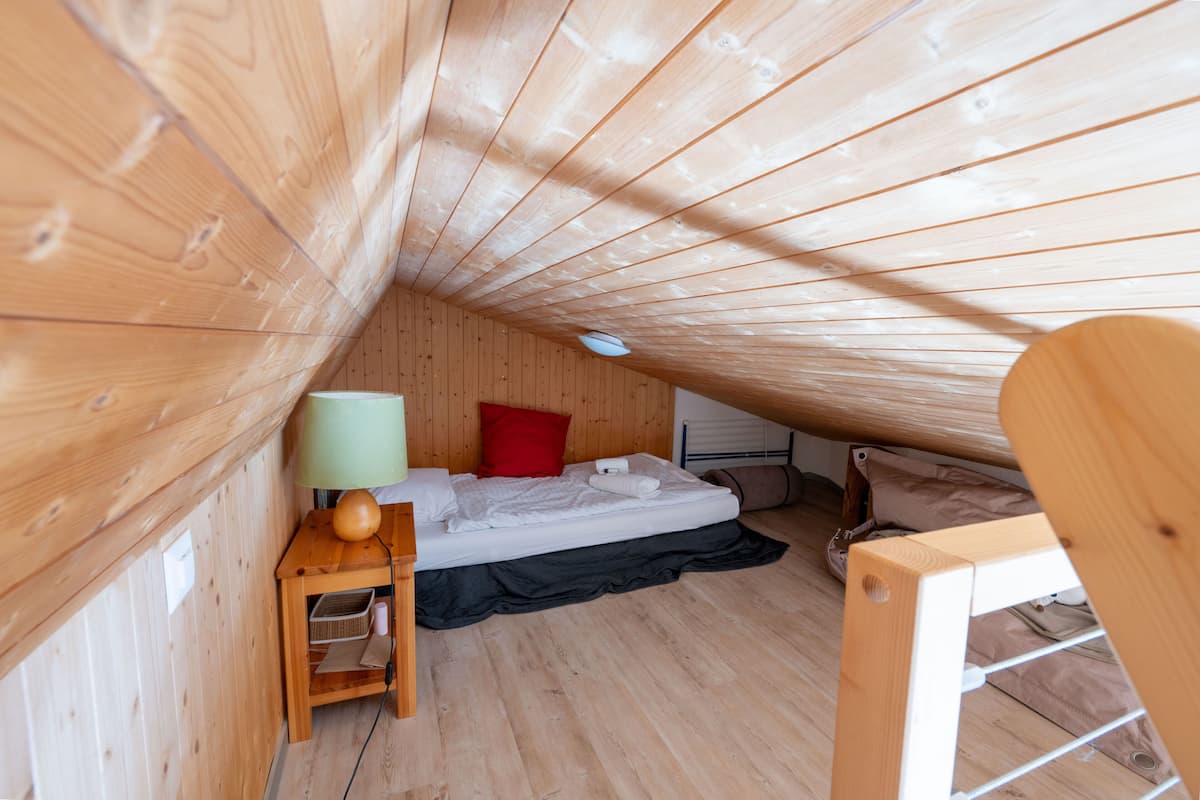 A small, wooden room with a sloped ceiling, a single bed, a nightstand with a lamp, and a window.