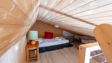 Ein kleiner Raum mit Holzwänden und schräger Decke, einem Einzelbett, einem Nachttisch mit Lampe und einem Fenster.