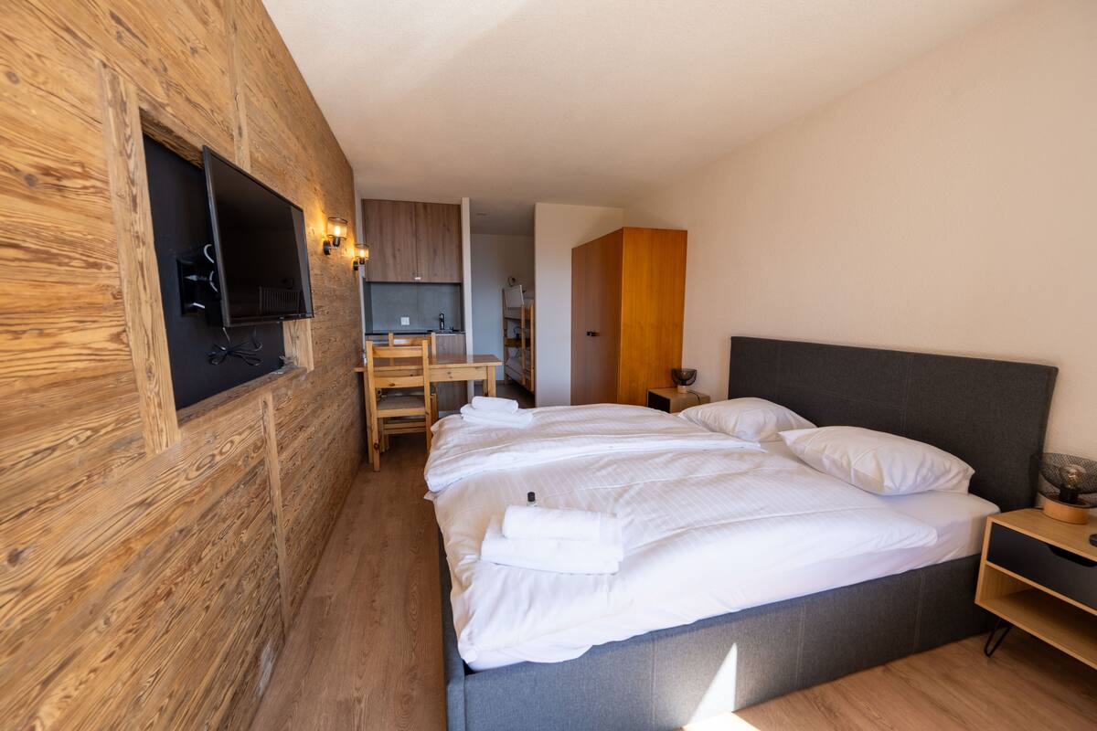 A modern hotel room with a wooden accent wall, a flat-screen TV mounted above a desk, a large bed with white linens, and a small bedside table.