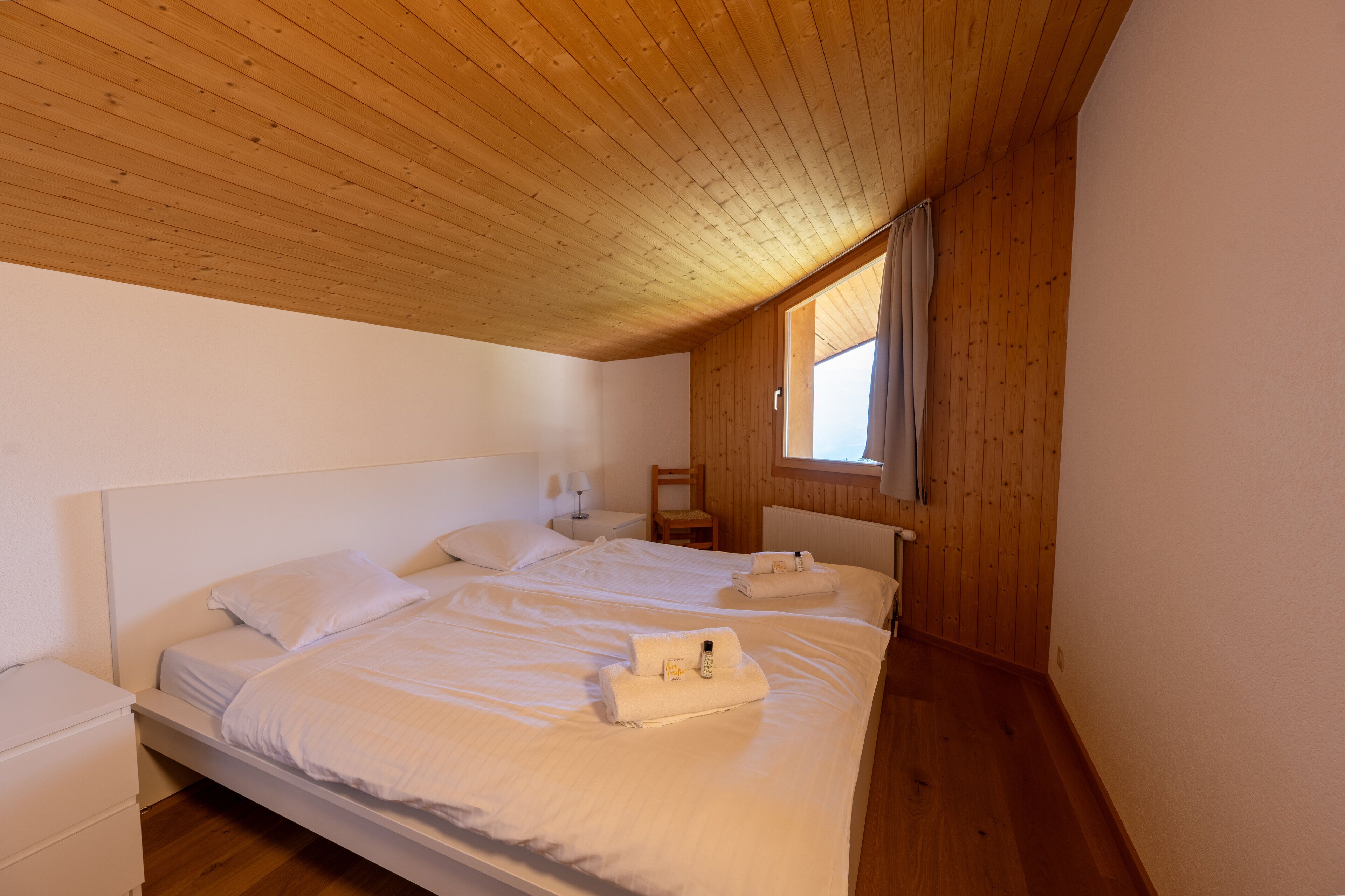 A bedroom with two beds, wooden ceiling, and a window with curtains.