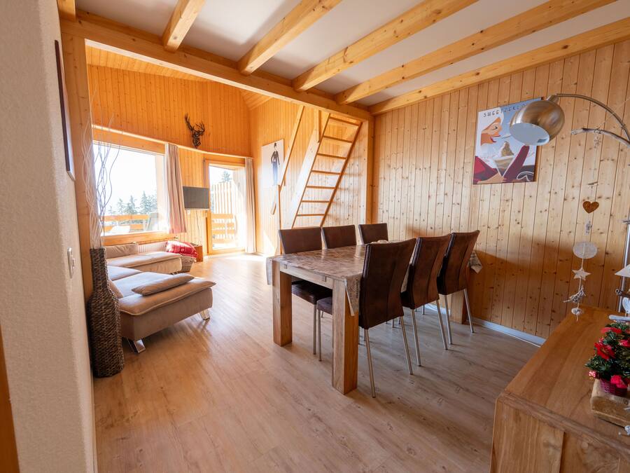 Een houten interieur met een eettafel en stoelen, een bank en een ladder tegen de muur.