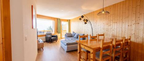 A spacious living area with a wooden dining table and chairs, a sofa, and a large window providing natural light.