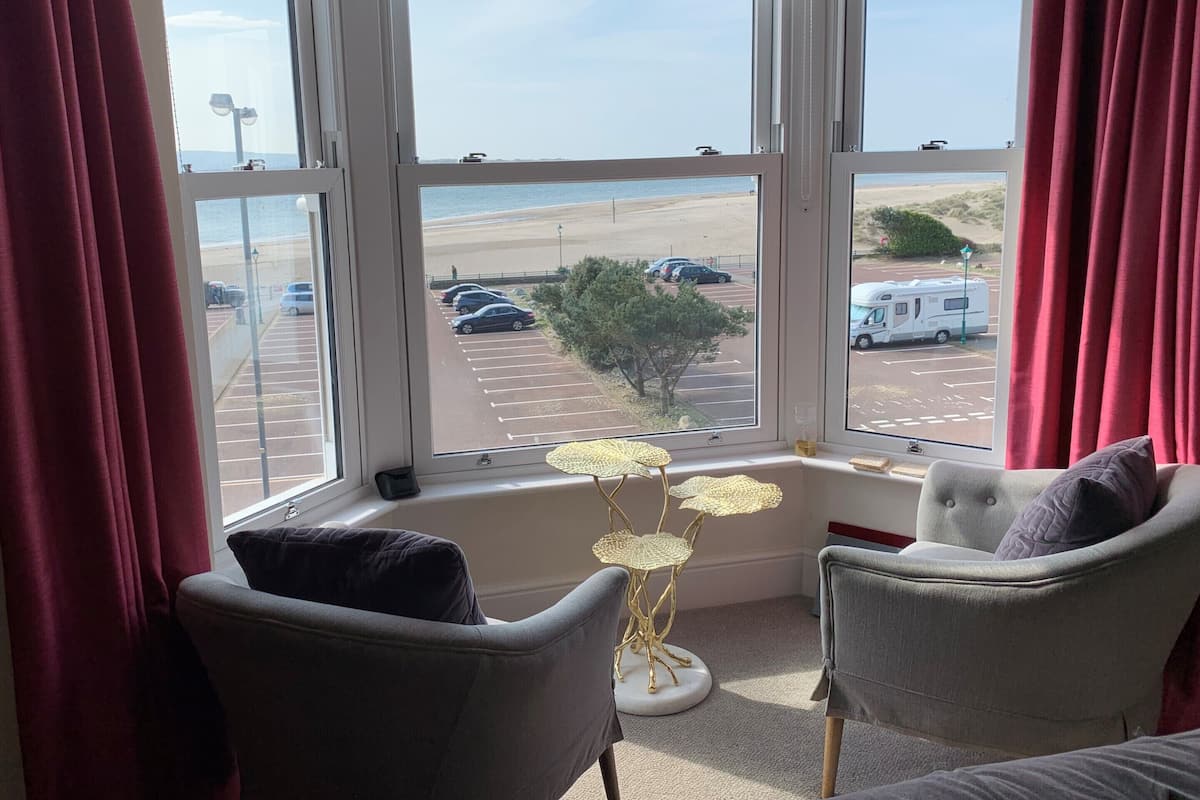 Quarto com uma grande janela com vista para uma praia e estacionamento, com duas cadeiras e uma pequena mesa.