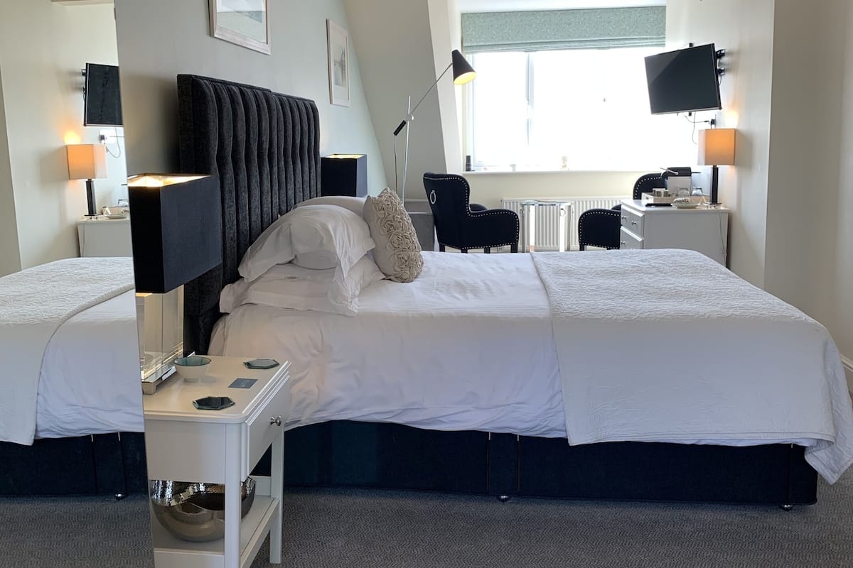 Quarto de hotel com uma cama grande, mesas de cabeceira, escrivaninha, cadeira, televisão e janela.