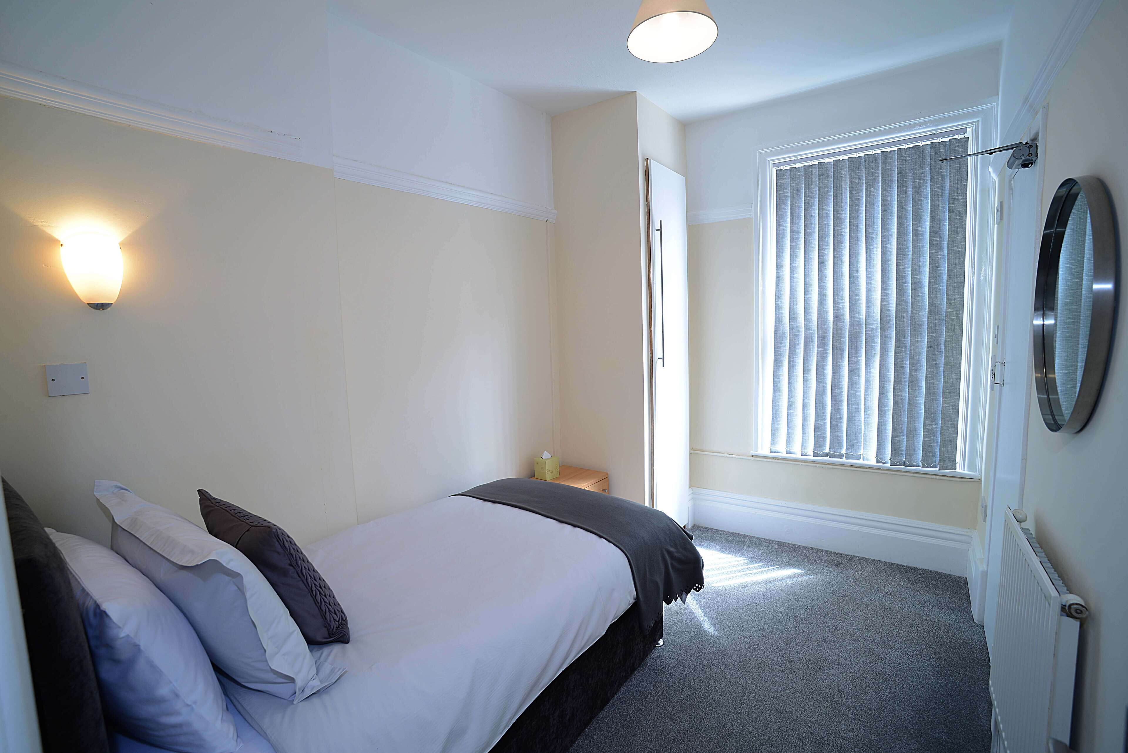 A bedroom with a bed, bedside tables, a wall-mounted light, a window with blinds, and a radiator.