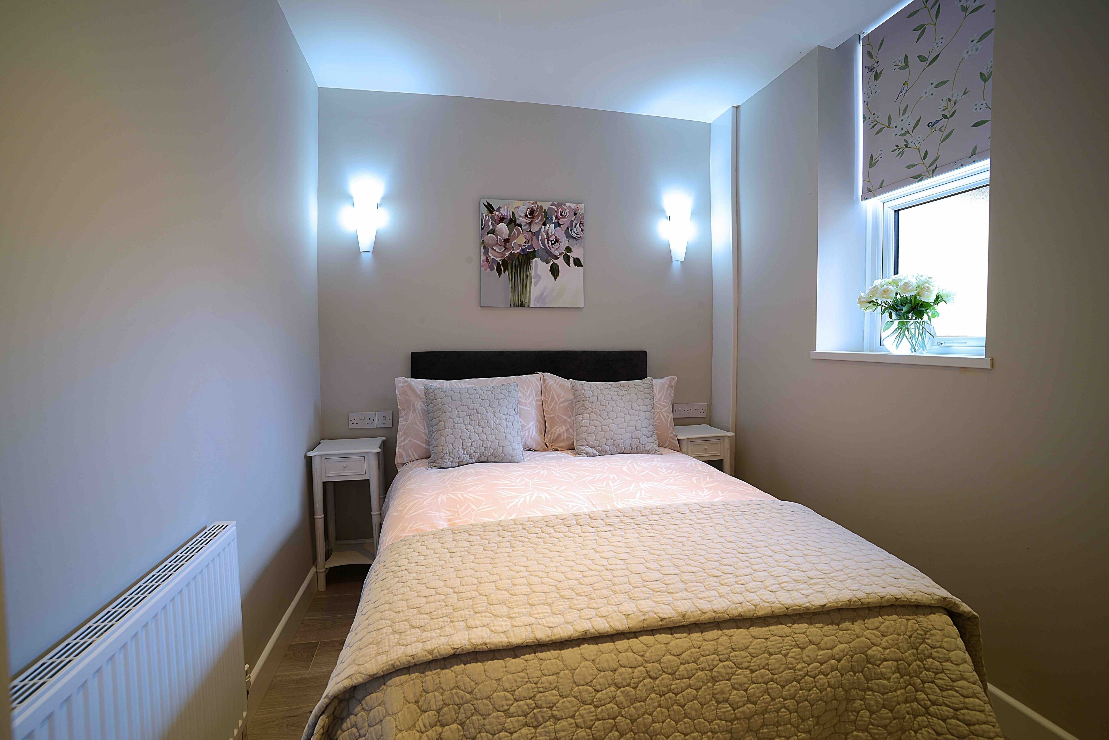 A bedroom with a bed, nightstands, a wall-mounted light, a window with a view, and a radiator.