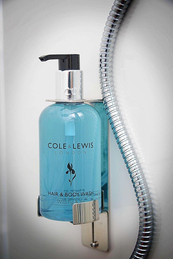 A bathroom with a blue bottle of Cole & Lewis hair and body wash mounted on a wall, next to a shower hose. - Twelve Holiday Apartments (Blackpool)