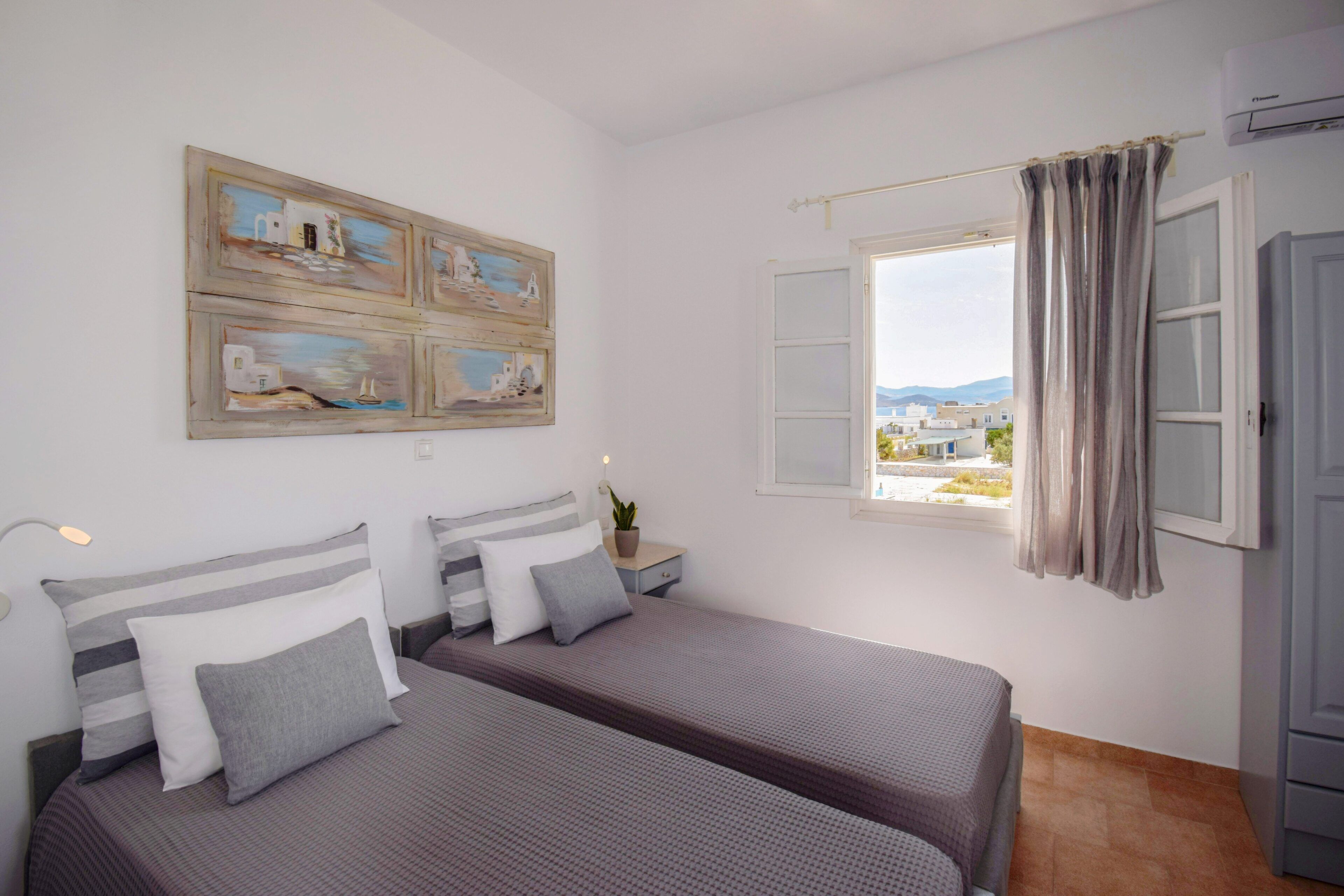A bedroom with two beds, a window with curtains, a wall-mounted artwork, and a bedside lamp.