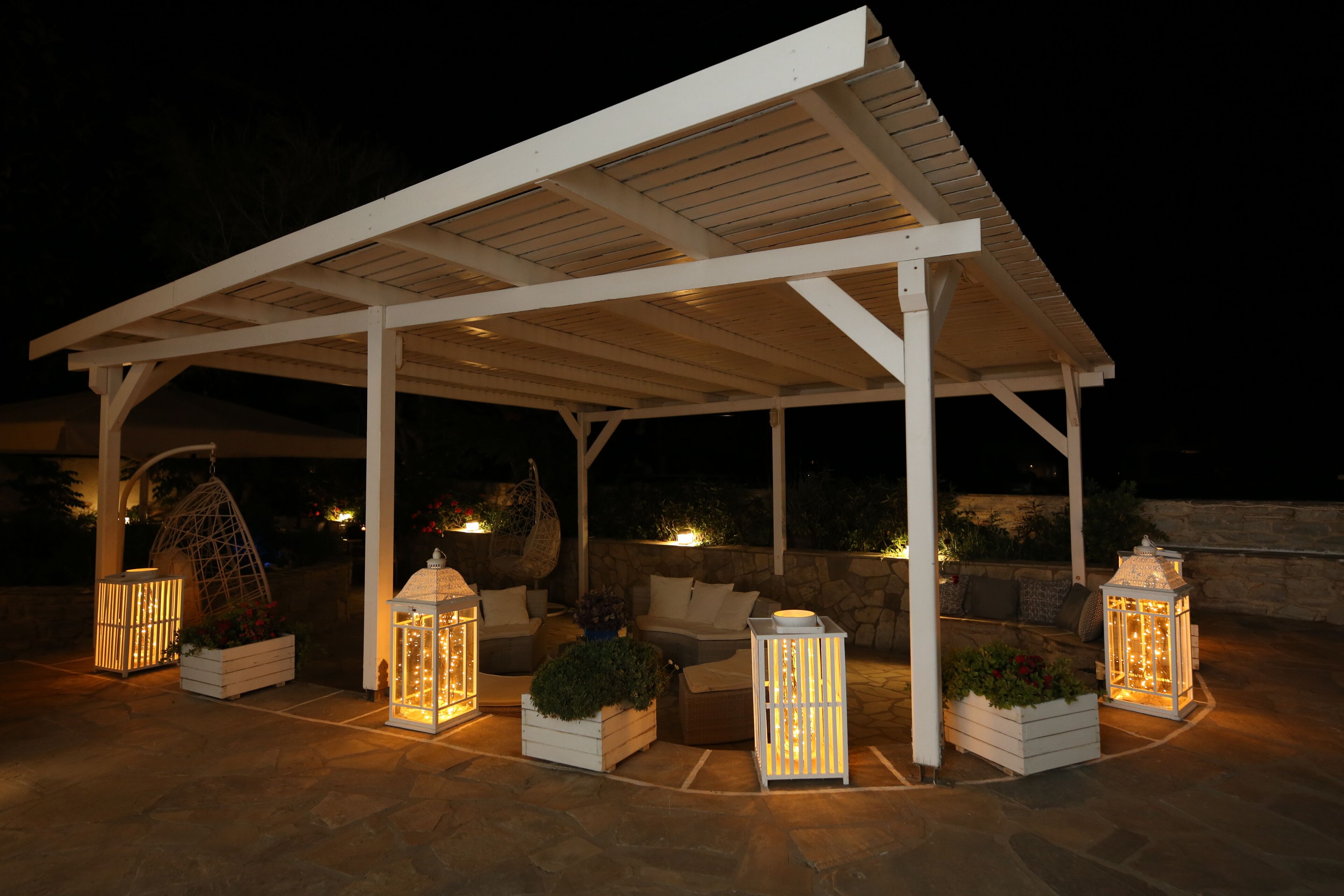A covered outdoor seating area with stone flooring, white wooden furniture, and illuminated lanterns.