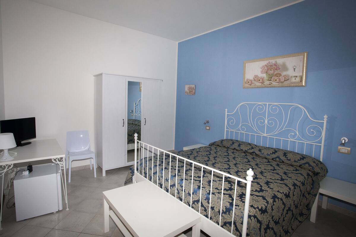 A bedroom with a bed, a desk with a chair, a small table, and a television.