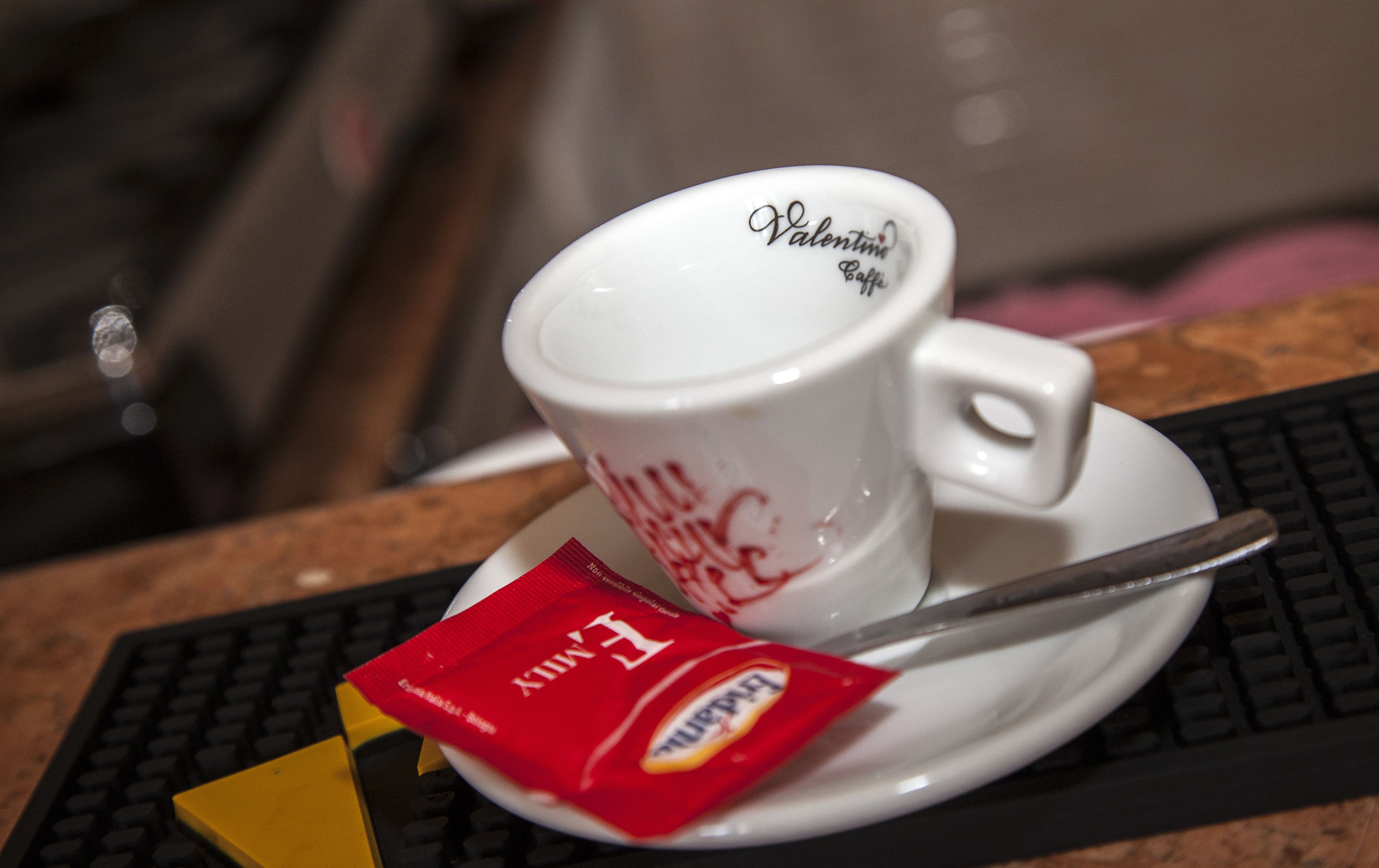 A white cup with "Valentina Cappuccino" written on it, placed on a saucer with a spoon. There is a packet of "Earl Grey" tea and a yellow napkin on the table.