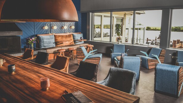 A modern lounge area with a wooden bar, comfortable seating, and large windows offering a view of the outdoors. - Hotel Zwartewater (Zwartsluis)