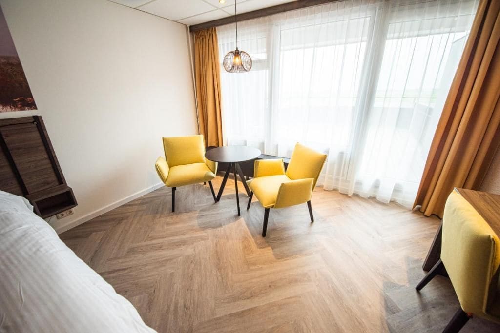 Una habitación de hotel con una cama, una mesa redonda con cuatro sillas amarillas y un ventanal con cortinas.