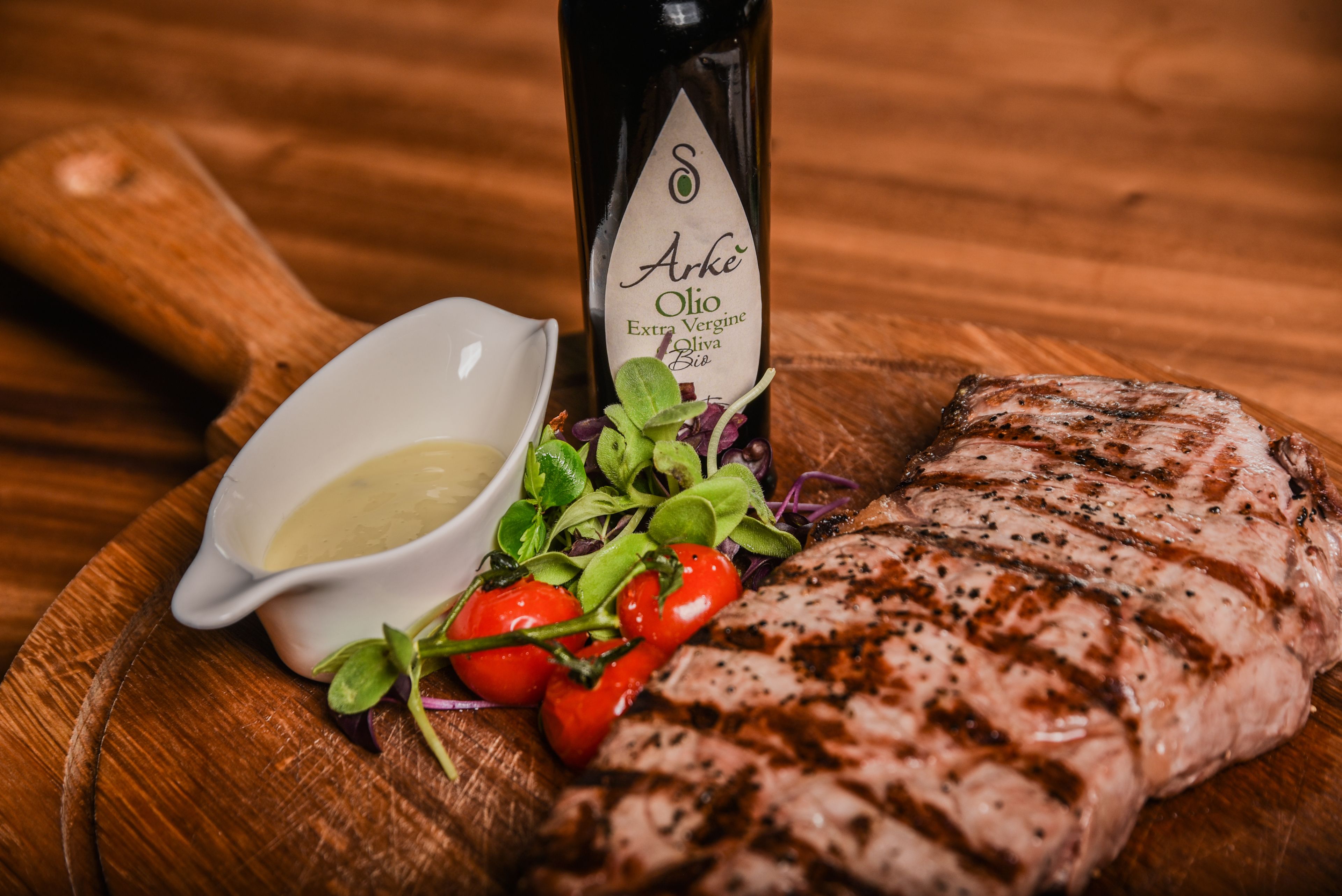 A grilled steak with a side of greens and cherry tomatoes, accompanied by a bottle of Arke Olio extra virgin olive oil.