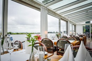 A restaurant with tables set for dining, large windows, and a view of the water. - Hotel Zwartewater (Zwartsluis)