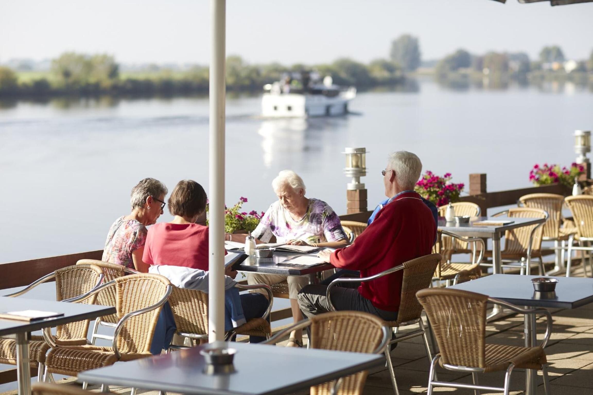 A group of people seated at tables on a deck overlooking a body of water, with a boat visible in the distance.