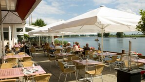 An outdoor café with wicker chairs and tables, white umbrellas, and a view of a lake with boats. - Hotel Zwartewater (Zwartsluis)