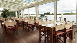A covered outdoor dining area with wooden tables and chairs, large windows overlooking a body of water and a boat. - Hotel Zwartewater (Zwartsluis)