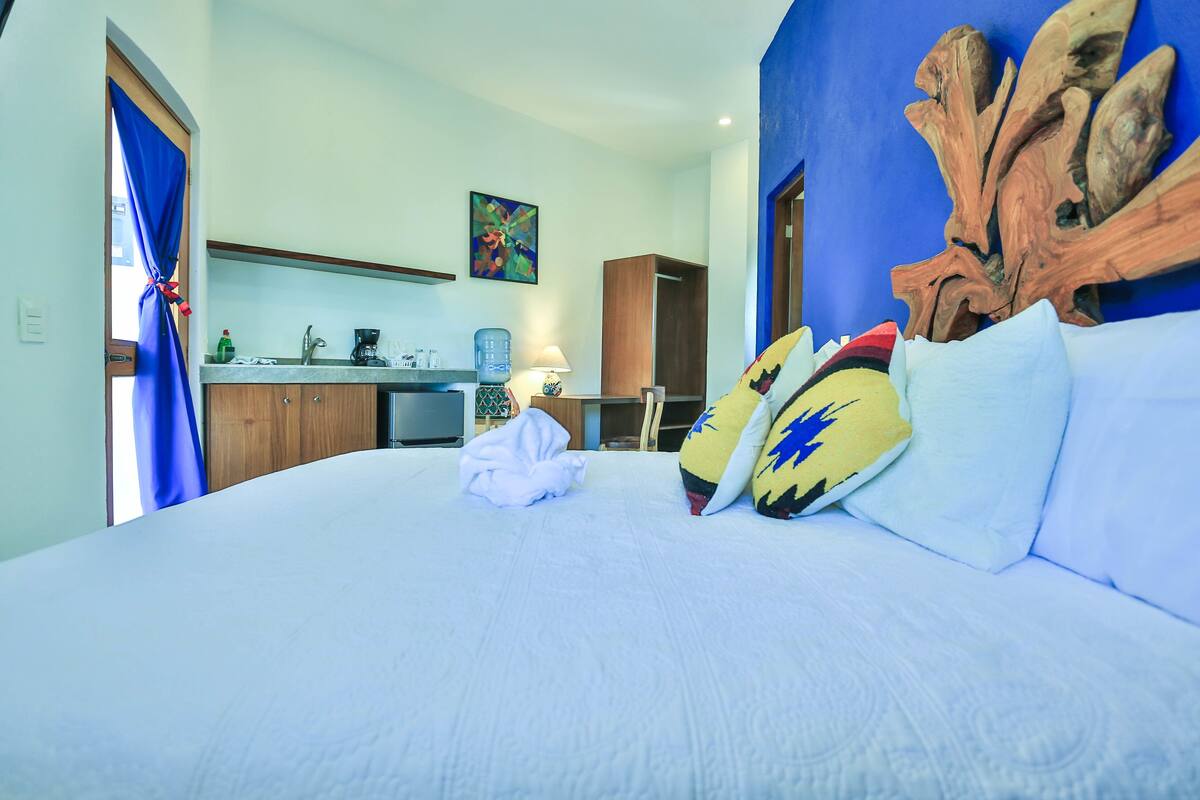 Ein Hotelzimmer mit Bett, Holz-Kopfteil, einem Gemälde, einer Küchenzeile und einem blauen Vorhang.