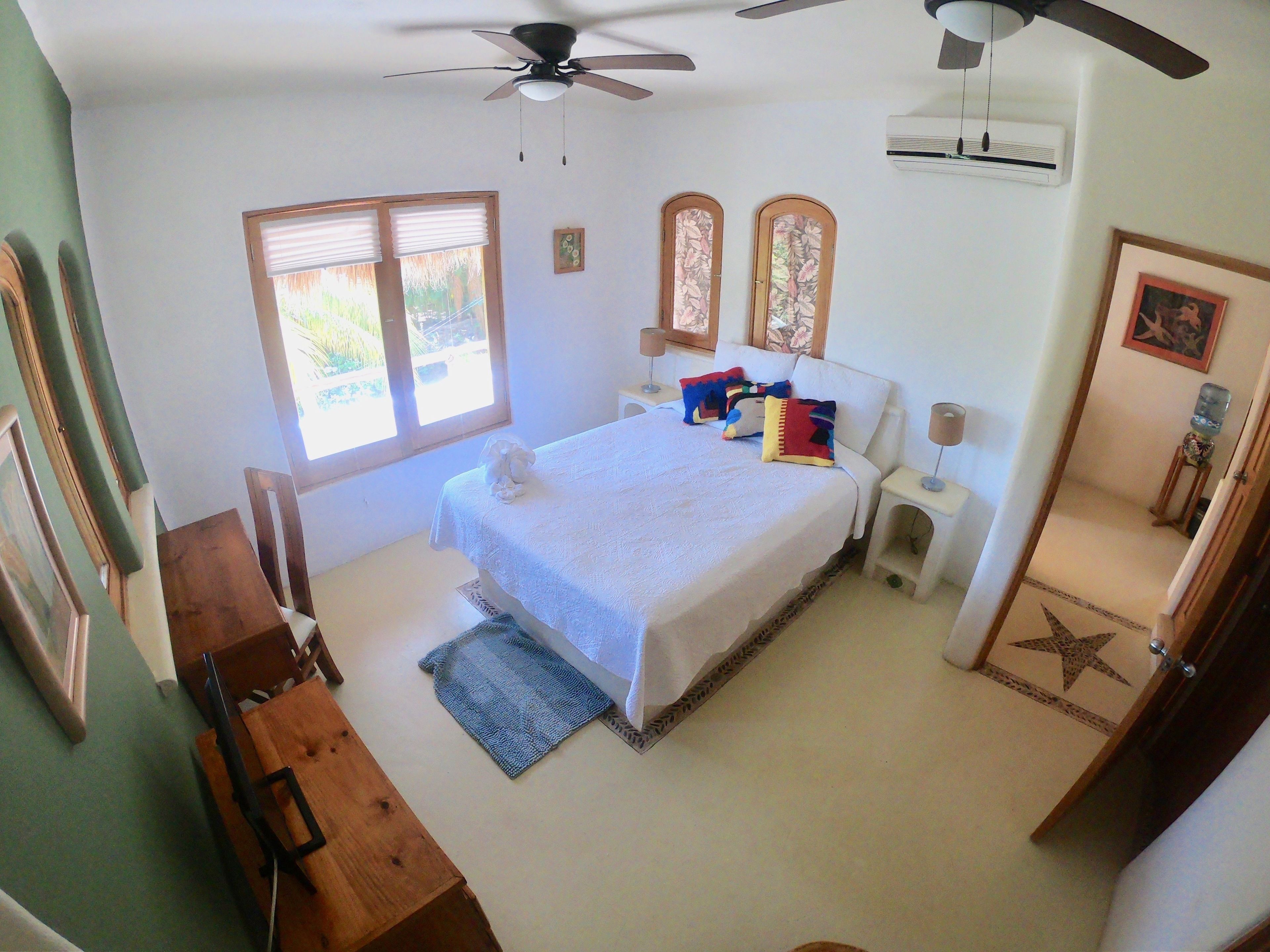 A bedroom with a bed, two nightstands, a ceiling fan, an air conditioner, a window with blinds, and a door with a star design.