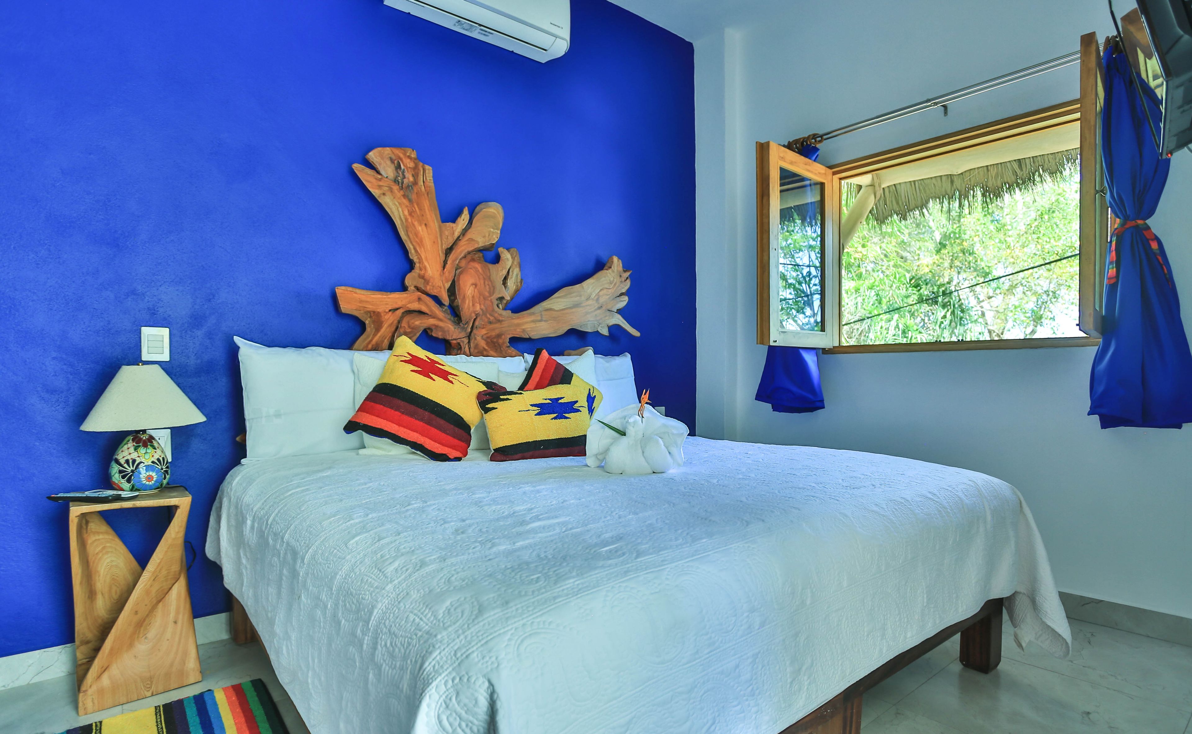 Un dormitorio con pared azul, cabecera de madera, ropa de cama blanca y una ventana con cortinas.