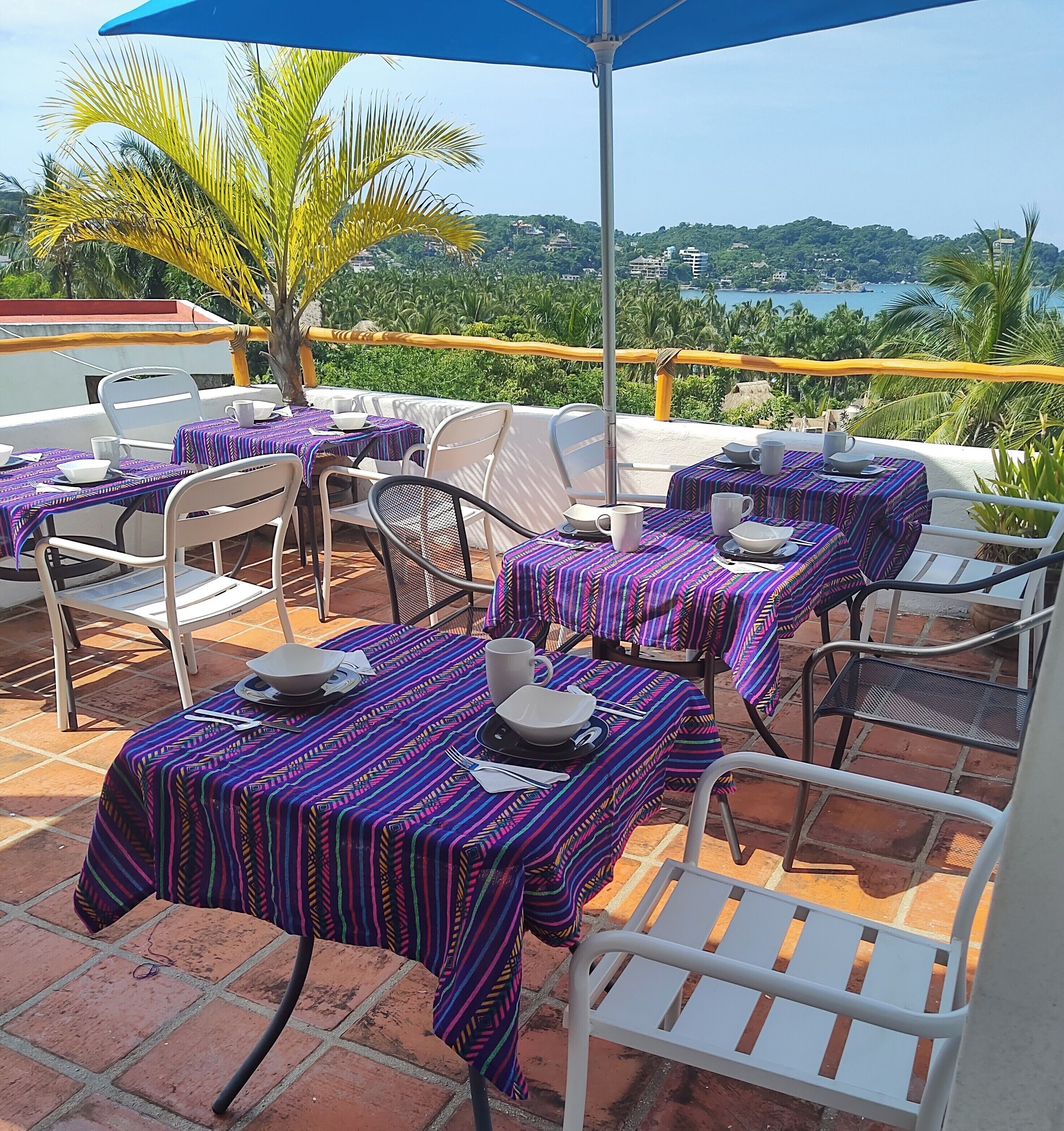 An outdoor dining area with tables covered in patterned tablecloths, white chairs, and a blue umbrella.
